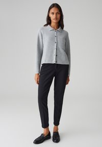 Cardigan a coste grigio chiaro con colletto, bottoni e taschino sul petto, abbinato a pantaloni neri aderenti e mocassini neri.