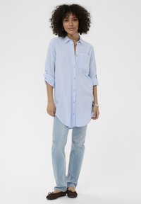 Vaaleansininen nappipaita, jossa on rintatasku, pitkät hihat käärittynä, yhdistettynä vaaleansinisiin farkkuihin ja ruskeisiin litteisiin kenkiin. Yksivärinen tausta.