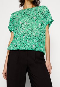 Blusa floral verde con mangas cortas, que presenta un corte holgado y patrones florales en blanco y negro. Combinada con pantalones negros.