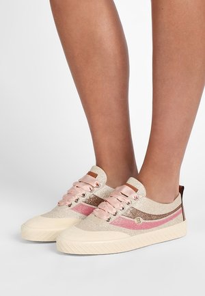 Pieds chaussés de baskets beige pailletées avec des accents roses et marron, des lacets en satin rose, et des semelles blanches texturées sur un fond blanc.