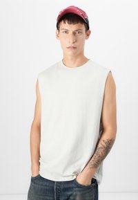 Lichtgrijze mouwloze shirt met een ronde hals, gecombineerd met een rood en zwart pet. De persoon heeft een tattoo op hun arm.