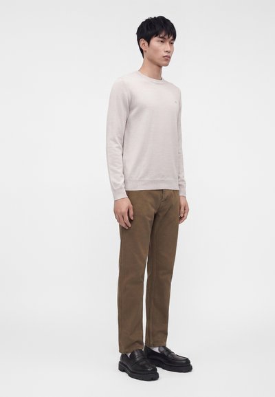 Jeune homme debout portant un pull beige clair à manches longues, un pantalon marron et des mocassins noirs sur un fond blanc uni.