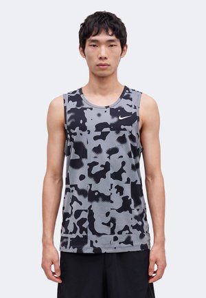 Tânăr purtând un maiou Nike camuflaj gri și negru fără mâneci, stând în fața unui fundal simplu gri deschis.