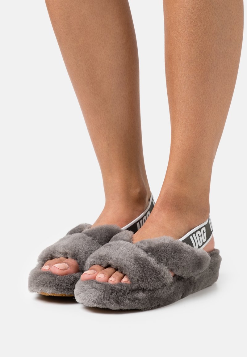 Pieds portant des sandales grises duveteuses avec des brides croisées et des bandes élastiques de talon noires et blanches de marque sur fond blanc.