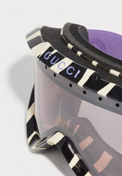 Occhiali da sci con un design a strisce nere e crema, lente sfumata e imbottitura in schiuma viola testurizzata. Caratterizzati da logo "GUCCI" impresso.