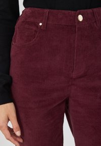 Bordeaux corduroybroek met verticale ribbels, voorzien van een voorkeux en een gouden knoopsluiting. Zachte textuur en aansluitende pasvorm zijn zichtbaar.