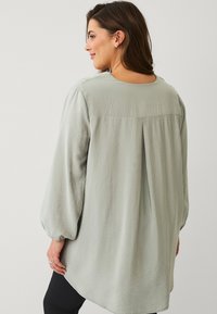 Lichtgroene blouse met lange, puffed mouwen. Heeft een soepele textuur en plooidetail aan de achterkant, met een ronde halslijn.
