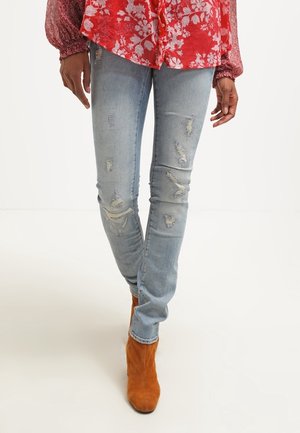 Personne portant un jean skinny déchiré bleu clair, un chemisier rouge à fleurs et des bottines en daim marron, debout devant un fond blanc uni.