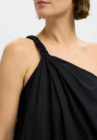 Top asimétrico negro con tirante trenzado y tejido drapeado, que presenta una textura suave y un corte holgado.