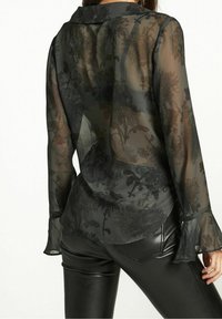 Transparente schwarze Bluse mit floralem Muster, langen Ärmeln, Knopfleistenkragen und Schlagmanschetten, kombiniert mit schwarzer, enger Hose.