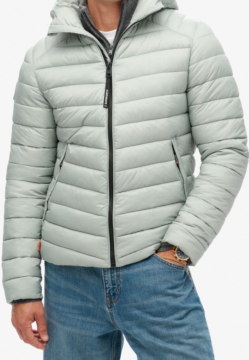 Chaqueta acolchada gris claro con capucha, con diseño acolchado horizontal, cremallera frontal y bolsillos laterales. Lleva puesta sobre una sudadera oscura.