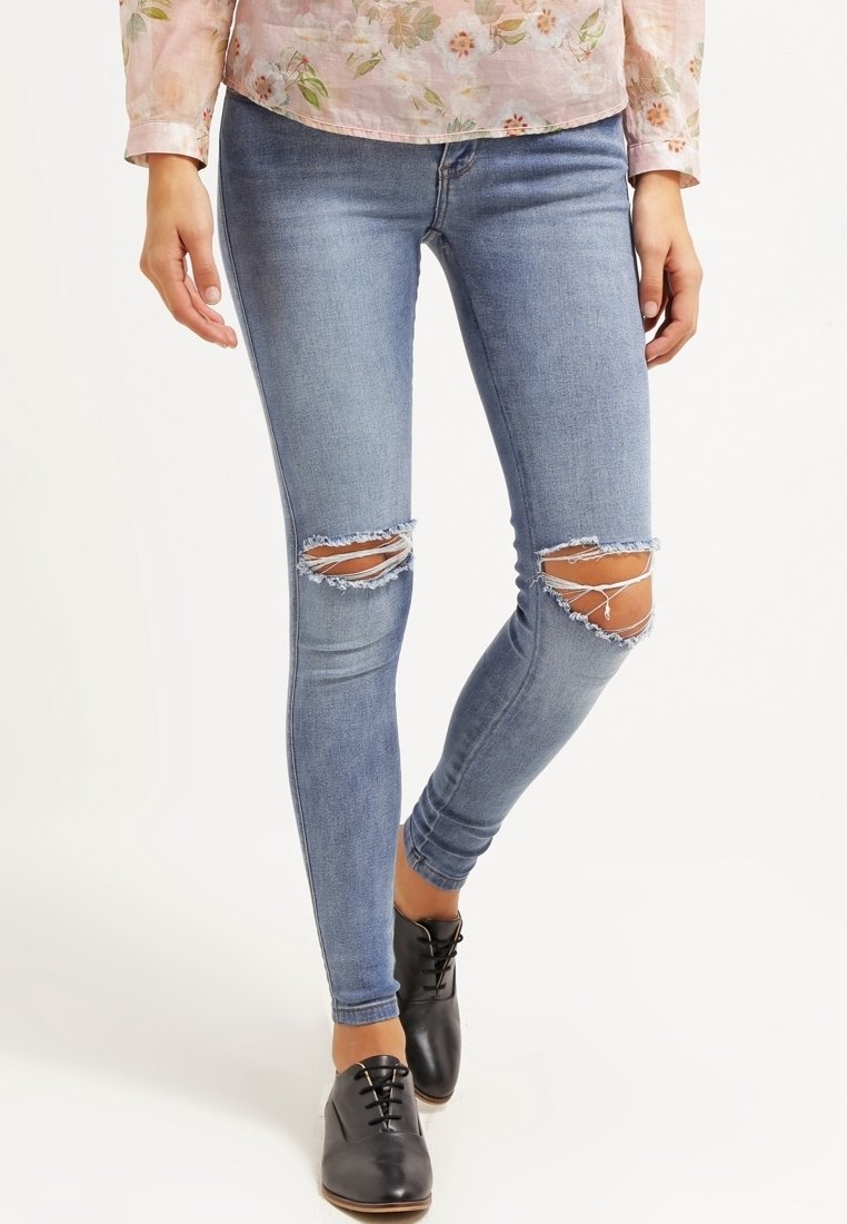 Vaqueros pitillo de mezclilla azul claro con roturas desgastadas en las rodillas y bordes deshilachados, complementados con zapatos negros con cordones.