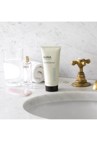 Máscara de crema hidratante en un tubo beige, botella de spray transparente, vaso de agua, herramienta de cuarzo rosa, toalla blanca, grifo de bronce sobre una superficie de mármol.