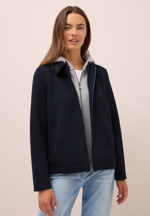 Femme portant une veste navy sur un sweat à capuche gris zippé et un jean bleu clair, se tenant contre un fond beige clair uni.