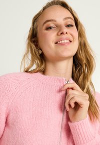 Sweat à zip rose en matériau doux et texturé, avec un col et des poignets côtelés. Détail de fermeture éclair argentée à l'avant pour un intérêt visuel.