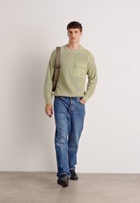 Un pull côtelé vert clair avec une poche avant, associé à un jean en denim bleu. La tenue est complétée par des chaussures noires et une bandoulière grise.