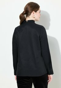 Zwarte langwerpige top met een hoge hals en een split op de rug. Soepele textuur, aansluitend ontwerp en met knopen bij de mouwen, gecombineerd met donkere broeken.