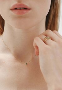 Collar de cadena de oro con un pequeño colgante de piedra preciosa verde. La modelo toca la cadena, mostrando un diseño minimalista contra la piel desnuda.