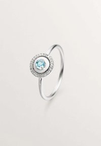 Anillo de plata con una piedra central azul claro rodeada de un arreglo circular de pequeñas piedras claras, con una banda lisa y pulida.