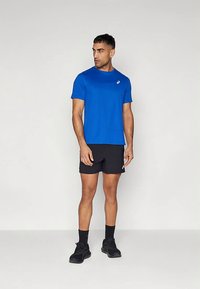 Blå kortermet treningsskjorte laget av glatt stoff, kombinert med svarte shorts. Har en logo på venstre bryst på skjorten.