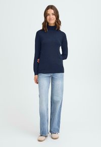 Marinblå polotröja, mjuk textur, figurnära design, kombinerad med ljusblå vida jeans. Skorna är beige med en enkel design.