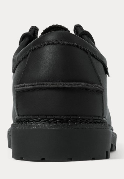 Toc pantof negru din piele, cu talpă groasă și cusături vizibile, văzut din spate pe un fundal deschis.