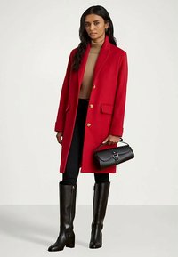 Γυναίκα φορώντας ένα κόκκινο παλτό, μπεζ ζιβάνγκο, μαύρα παντελόνια, μαύρες μπότες ως το γόνατο, κρατώντας μια μαύρη τσάντα με πόρπη.