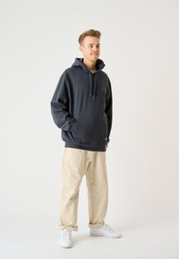 Dunkelgrauer Hoodie mit einer vorderen Tasche und Kordelzug, kombiniert mit beigen Hosen und weißen Sportschuhen. Der Hoodie hat ein kleines Logo auf der Brust.