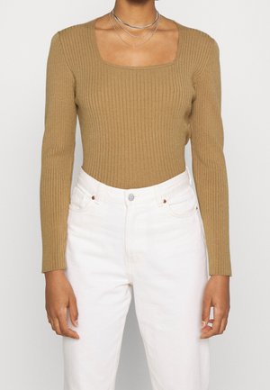 Persona que lleva una camiseta de manga larga color camello con textura acanalada, escote cuadrado y pantalones blancos de talle alto.
