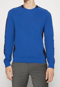 Maglione azzurro lavorato a maglia con scollo rotondo, dettagli neri sulle spalle e logo sul petto. Maniche e orlo a coste.