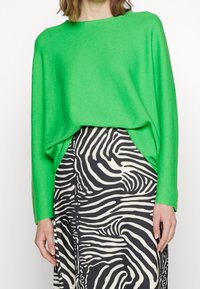 Pull vert vif surdimensionné à manches longues, en matériau doux, associé à une jupe à rayures zébrées noires et blanches.