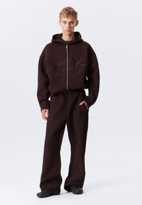 Brun zip-up hettegenser og matchende joggebukser, begge laget av tykk stoff. Hettegenseren har et embossert design og kengurulomme.