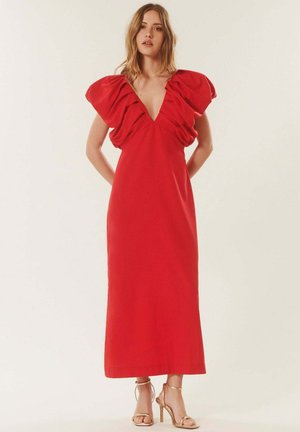 Femme aux cheveux ondulés portant une robe rouge longue jusqu'au sol avec de grandes manches froufroutantes et un décolleté en V profond, debout les mains derrière le dos.