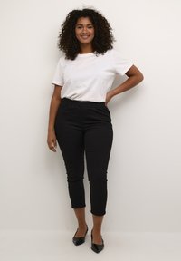 Pantaloni neri a metà gamba con vita elasticizzata, abbinati a una maglietta bianca semplice. Il modello sta in piedi con una mano in vita, sorridendo.
