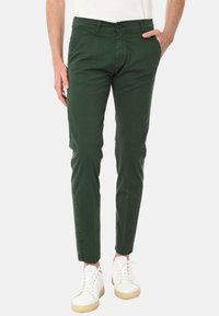 Chinos verdi slim-fit con una texture liscia, dotati di chiusura a bottone e tasche laterali, abbinati a sneakers bianche e una maglietta leggera.