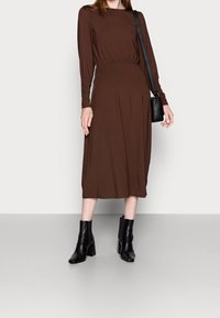 Brun långärmad klänning med en samlad midja, flödande kjol och rund halsringning, matchad med svarta blockklack stövletter.