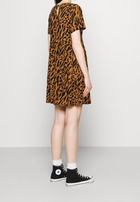 Person som bär en kortärmad klänning med brun och svart djurmönster, svarta hög-top sneakers och vita strumpor, som står mot en enhetlig bakgrund.