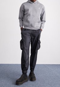 Felpa grigia con cappuccio e tasca a marsupio, abbinata a pantaloni cargo neri con tasche a zip e polsini elasticizzati, e stivali neri con tacco a piattaforma.