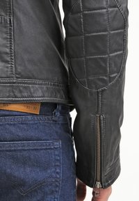 Nahaufnahme eines ärmels einer schwarzen Lederjacke mit gepolstertem Ellbogen und Reißverschluss, getragen mit blauen Jeans, die ein braunes Ledergröße-Etikett zeigen.