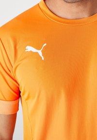 Orange idrottströja i slätt tyg, med rund halsringning, korta ärmar och meshdetaljer. Vit logotyp på bröstet.