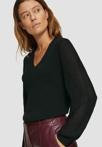 Pull noir avec un col en V, corps ajusté et manches transparentes rayées. Porté avec un pantalon en cuir bourgogne taille haute. Texture lisse.