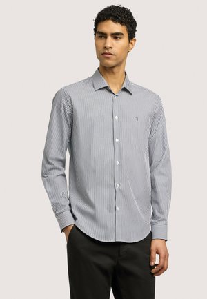 Mężczyzna ubrany w białą i czarną koszulę w pionowe paski z długim rękawem oraz czarne spodnie, stojący z jedną ręką w kieszeni.