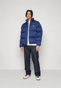 Ναυτικό puffer μπουφάν με όρθιο γιακά, μπροστινό κλείσιμο με φερμουάρ και λεπτομέρεια λογότυπου στο αριστερό στήθος, συνδυασμένο με σκούρες τζιν παντελόνες και λευκά αθλητικά παπούτσια.