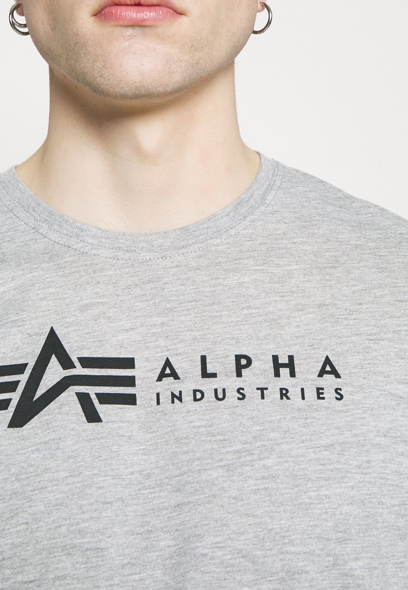 Camiseta de algodón gris con cuello redondo. Presenta un logo negro con el texto "ALPHA INDUSTRIES" destacado. Textura suave.
