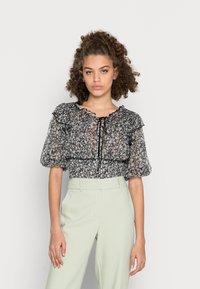 Blusa transparente preta com flores, mangas bufantes, acabamento em babados e cintura franzida. Combinada com calças sob medida verde claro para um visual contrastante.