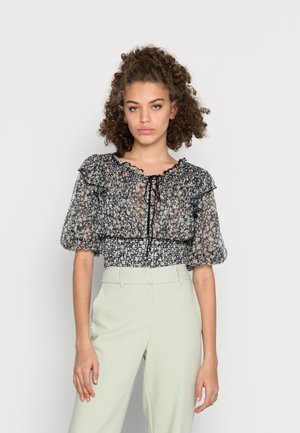 Blusa transparente preta com flores, mangas bufantes, acabamento em babados e cintura franzida. Combinada com calças sob medida verde claro para um visual contrastante.