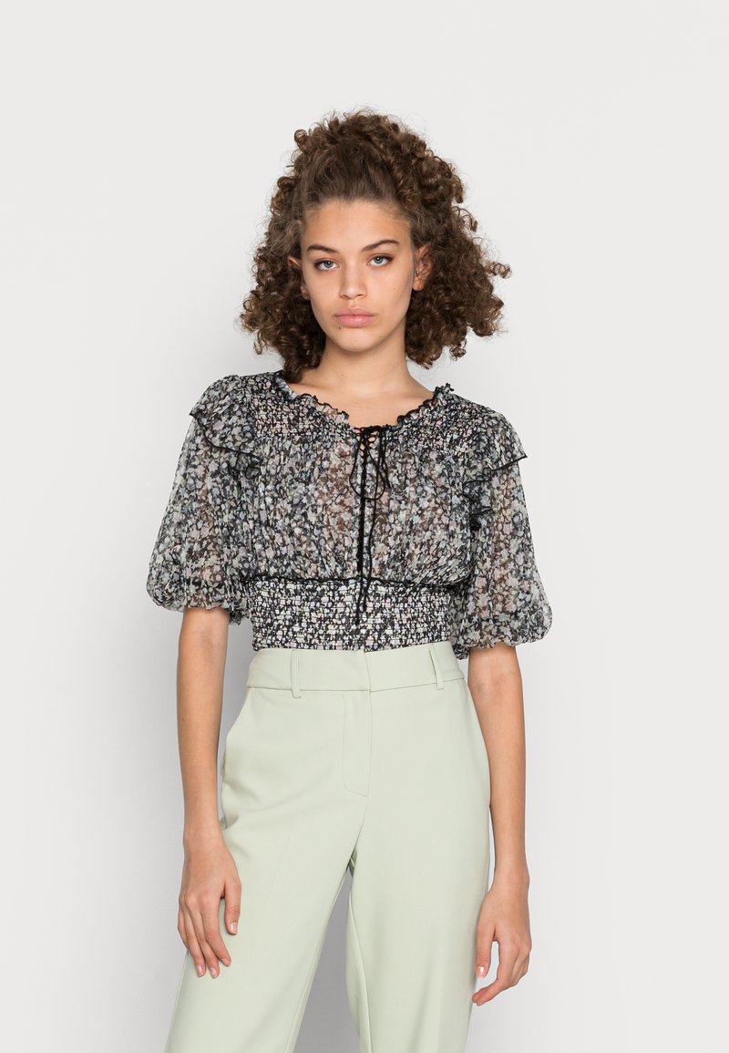 Blusa transparente preta com flores, mangas bufantes, acabamento em babados e cintura franzida. Combinada com calças sob medida verde claro para um visual contrastante.