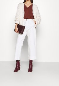 Pantalons blancs à jambes larges avec des ourlets retroussés, associés à un haut en V marron et un cardigan texturé crème, tenant un sac pochette bordeaux.