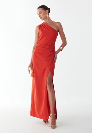 Žena u narančastoj haljini bez rukava s jednim ramenom i prorezom na bedrima, zlatnim naušnicama, narukvicom, torbicom i bež sandalama s visokim potpeticama na bijeloj pozadini.