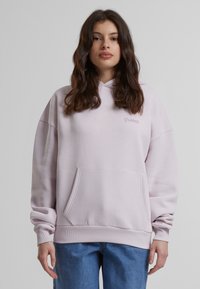 Hoodie oversize rose clair en tissu doux, avec une poche frontale, des poignets côtelés et un logo brodé discret sur la poitrine.